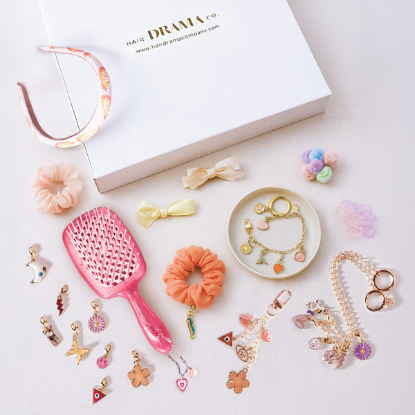Collection of hair accessories including a pink brush, scrunchies, and decorative hair ties on a white surface with 'Hair Drama Co' branding.