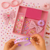 A child's hands are shown crafting a hair accessory with items from a pink and lilac DIY hair drama box, which includes various charms, crystals, hair bands, scrunchies, and bows.
