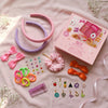 A child's hands are shown crafting a hair accessory with items from a pink and lilac DIY hair drama box, which includes various charms, crystals, hair bands, scrunchies, and bows.