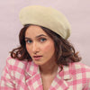 A woman wearing a beige knitted wool beret with pearl embellishment on a pink checkered coat, against a pink background.