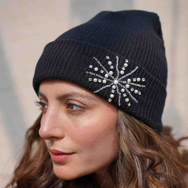 A woman wearing a black ribbed beanie with embellishments of pearls, crystals, and a camera charm.