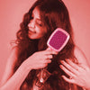 Woman brushing her hair with a pink brush against a pink background