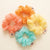 Four sheer chiffon hair scrunchies in peach, orange, yellow, and light blue, arranged in a circle on a warm off-white background. Each scrunchie has a small fabric tag with the brand name 'IICQ' printed on it.