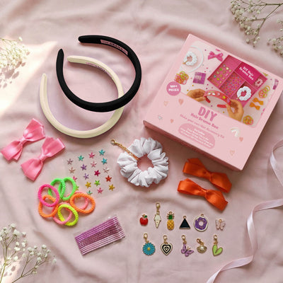 A selection of hair accessories including charms, crystals, and pom poms arranged neatly in a pink box, with a person holding a hair band decorated with charms.