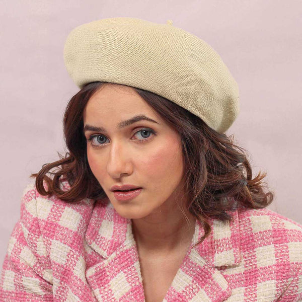 A woman wearing a beige knitted wool beret with pearl embellishment on a pink checkered coat, against a pink background.