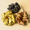 A set of three silk satin scrunchies in gold, bronze, and black colors, displayed on a neutral background.