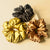 A set of three silk satin scrunchies in gold, bronze, and black colors, displayed on a neutral background.