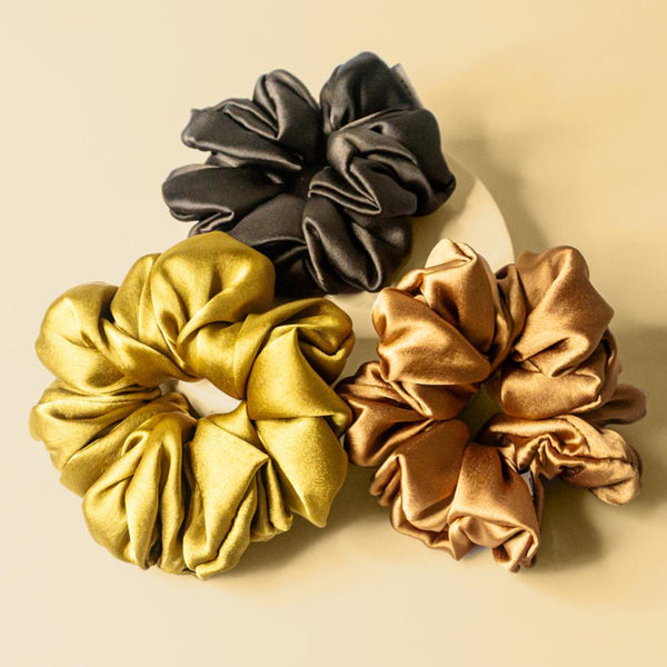 A set of three silk satin scrunchies in gold, bronze, and black colors, displayed on a neutral background.